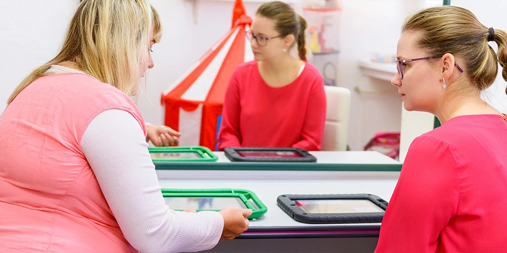 A speech therapist using a tablet device to communicate with a young woman in a therapy session, with both reflected in a mirror. 
