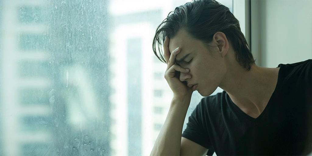 Young man sitting by a window with his hand on his face, appearing overwhelmed or anxious. 