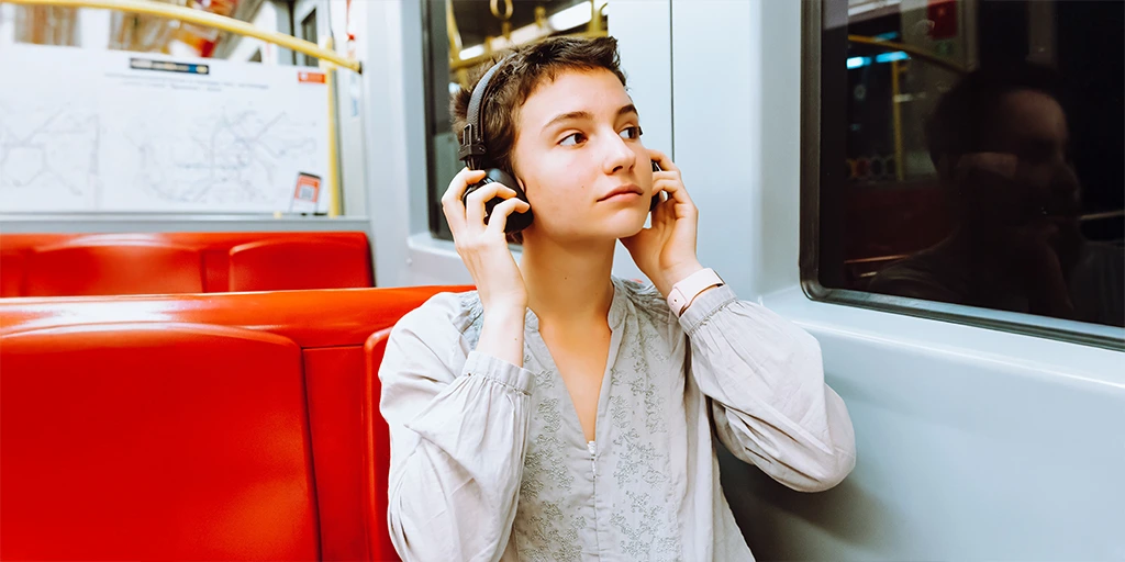 A young woman with short hair wearing headphones and looking out the window while riding public transportation, possibly using an AAC device for communication support. 