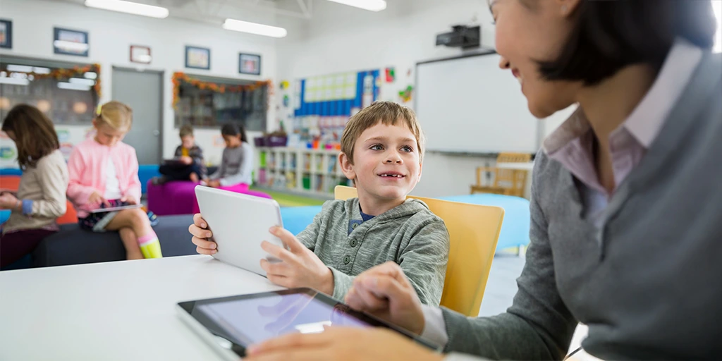 A young boy holding a tablet and smiling while interacting with a teacher or therapist in a classroom setting, with other children using devices in the background. 