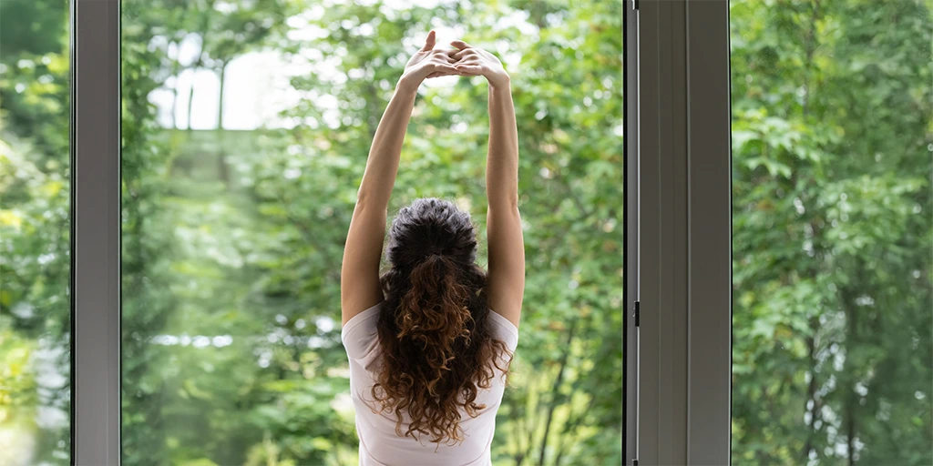 Woman stretching in front of a large window with a forest view, symbolizing a calm start to a sensory-friendly homeschool day. 