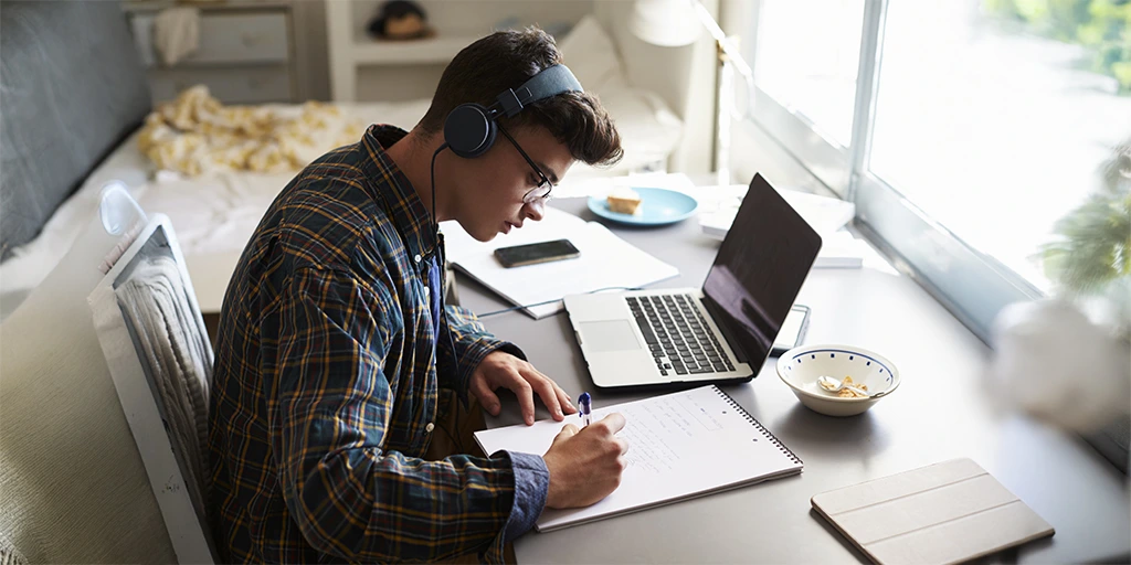 Teen boy with headphones studying at a tidy desk with natural light, using a laptop and writing in a notebook. 