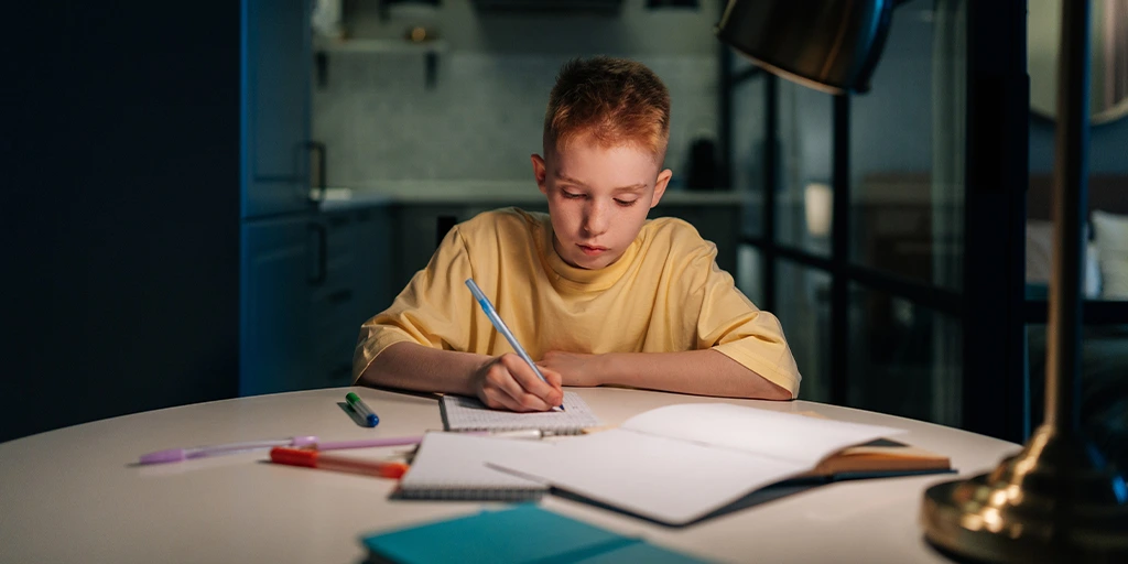 Young boy focused on homework at a round table under warm lighting, with notebooks and colored pens. 