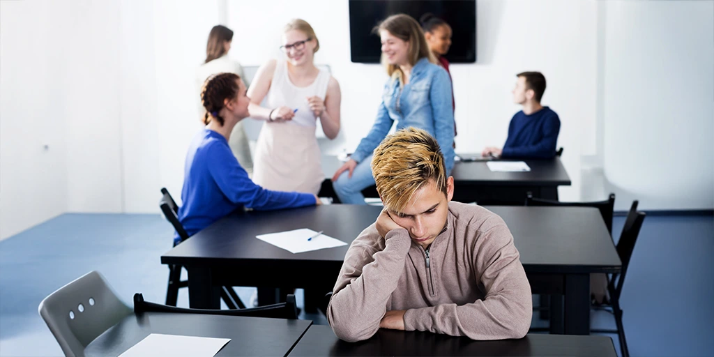 Teen boy sitting alone in a classroom while peers talk in the background, illustrating social exclusion often faced by autistic individuals.