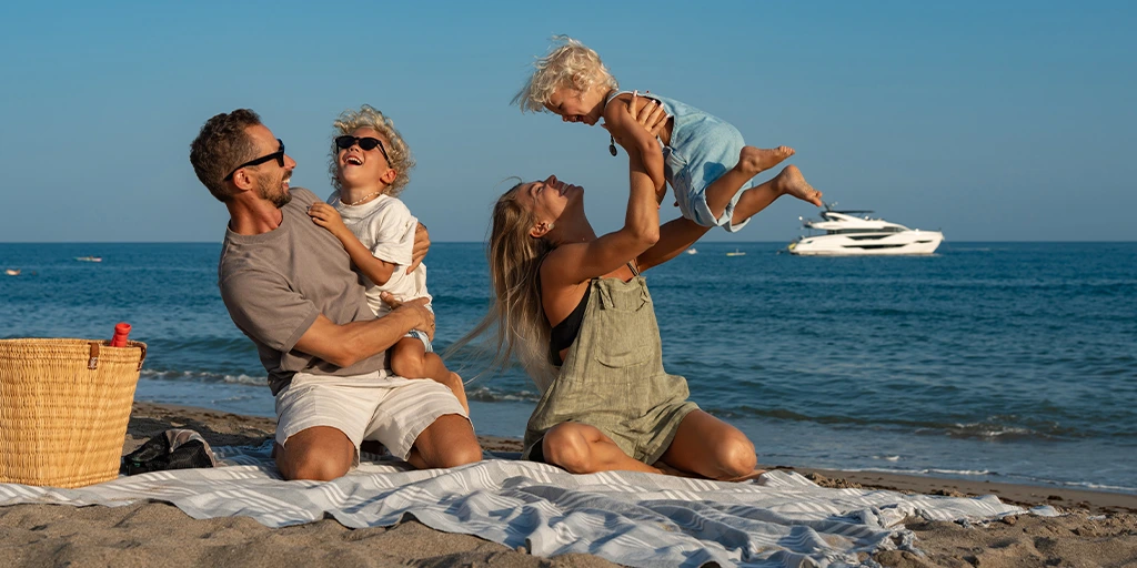 A joyful family enjoys a sunny day at the beach, with parents playing and laughing with their two young children near the water.