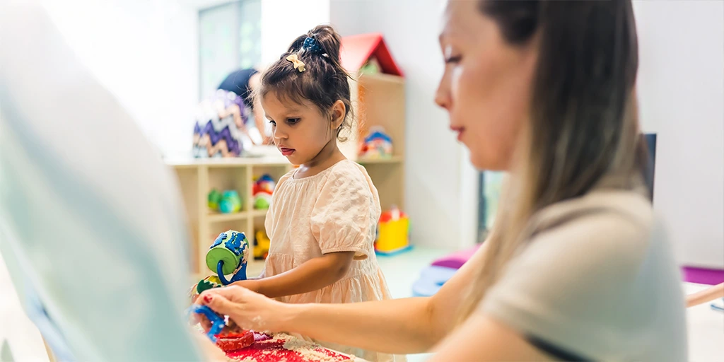 A young girl with a focused expression plays with colorful sensory toys next to an adult caregiver in a calm, indoor therapy or classroom setting. 