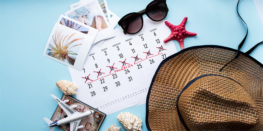 A travel-themed flat lay featuring a straw hat, sunglasses, calendar marked for vacation, seashells, and airplane model on a light blue background. 